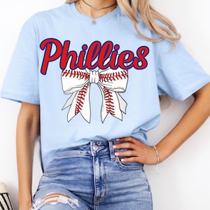 May include: A light blue t-shirt with the word "Phillies" in red and blue lettering. A white bow with red stitching and a baseball design is centered on the shirt.