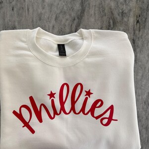 May include: White crew neck sweatshirt with the word "Phillies" in red script across the front. Two red stars flank the word. The sweatshirt is folded neatly on a gray surface.