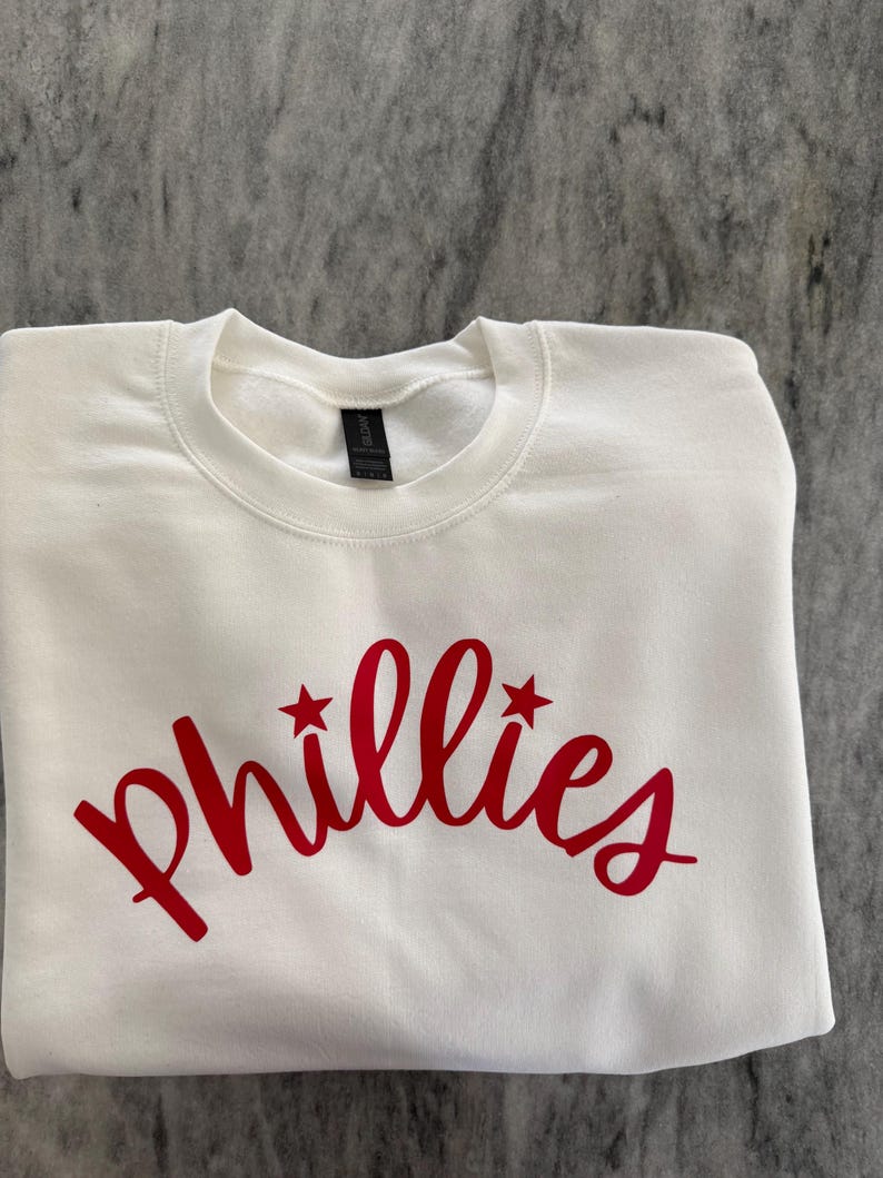 May include: White crew neck sweatshirt with the word "Phillies" in red script across the front. Two red stars flank the word. The sweatshirt is folded neatly on a gray surface.