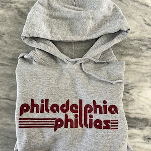 May include: A gray hooded sweatshirt with the text "philadelphia phillies" in maroon. The text is stylized with horizontal lines underneath. The sweatshirt is folded and has a drawstring at the hood.