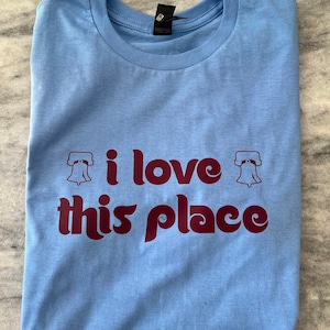 May include: A light blue t-shirt with a maroon graphic that says "i love this place" with a liberty bell graphic on each side of the text.