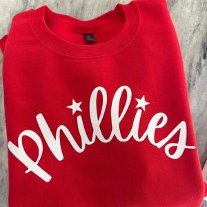 May include: A folded red sweatshirt with the word "Phillies" in white script across the front. Two white stars flank the word. The sweatshirt is made of a soft material.