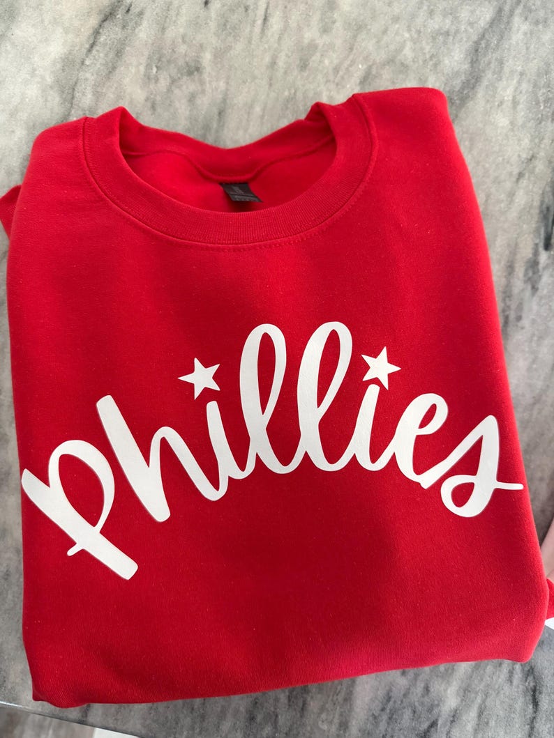 May include: A folded red sweatshirt with the word "Phillies" in white script across the front. Two white stars flank the word. The sweatshirt is made of a soft material.