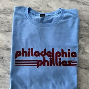 May include: A light blue t-shirt with the words "Philadelphia Phillies" printed in maroon. The shirt is folded neatly, showcasing the team's name in a stylized font with horizontal stripes.
