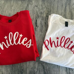 May include: Two shirts, one red and one white, each with the word "Phillies" in a script font with star accents. The red shirt is a crew neck sweatshirt, and the white shirt is a t-shirt.