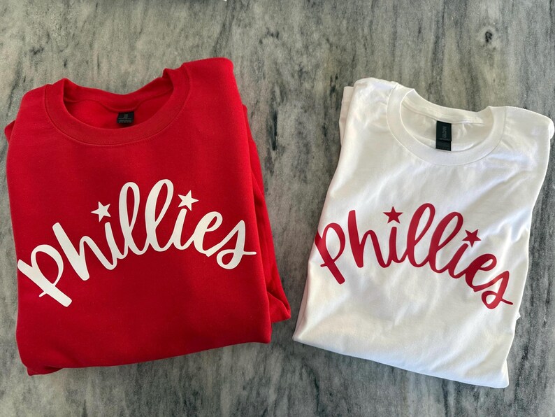 May include: Two shirts, one red and one white, each with the word "Phillies" in a script font with star accents. The red shirt is a crew neck sweatshirt, and the white shirt is a t-shirt.