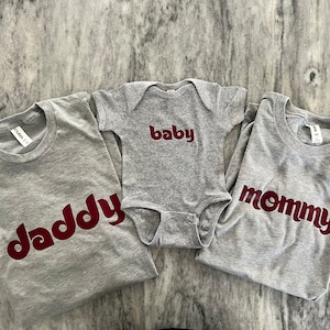 May include: Three grey t-shirts with the words "daddy", "mommy", and "baby" printed in maroon.