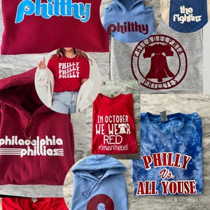 May include: Assortment of t-shirts and hoodies in various colors, including red, blue, and gray. The apparel features text and graphics related to Philadelphia, including "Phillies," "Philly," and "Ring That Jawn."