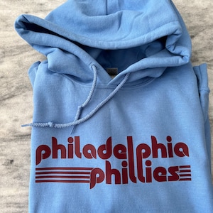 May include: A light blue hoodie with the words "Philadelphia Phillies" in maroon lettering across the front. The hoodie has a drawstring and a hood. The fabric appears to be a soft, comfortable material.