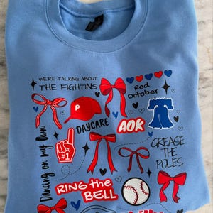 May include: Light blue sweatshirt with a variety of red, white, and blue graphics. The graphics include a baseball cap, a baseball, a bell, bows, and the words "We're talking about the Fightins", "Red October", "AOK", "Daycare", "Dancing on my own", "Ring the bell", "Grease the poles", and "Philly".