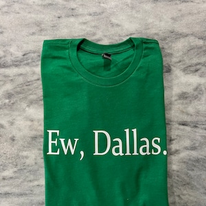 May include: A folded green t-shirt with the white text "Ew, Dallas." The shirt is made of a soft material and has a crew neck. The text is centered on the front of the shirt.