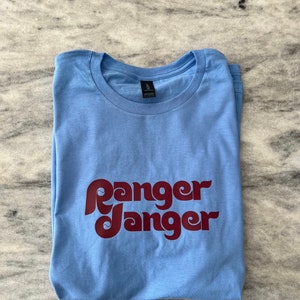 May include: A light blue t-shirt with a maroon "Ranger Danger" graphic printed on the front.