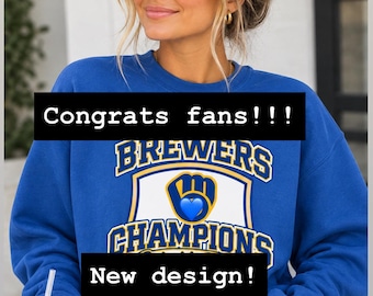 May include: A royal blue crewneck sweatshirt featuring a graphic with the text 'BREWERS' and 'CHAMPIONS' surrounding a baseball glove logo with a blue heart. The text 'Congrats fans!!!' and 'New design!' are overlaid on the sweatshirt.