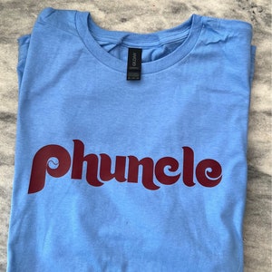May include: A light blue t-shirt with a maroon graphic that says "phuncle" in a retro font.