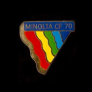 Puede incluir: Un pin triangular dorado con una pancarta azul que dice "MINOLTA CF 70". El pin presenta un diseño de arcoíris con rayas rojas, naranjas, amarillas, verdes, azules y moradas.