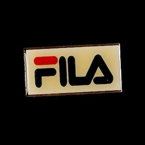 Peut inclure: Une épingle rectangulaire avec le mot "FILA" en lettres noires grasses. L'épingle a un fond crème avec une bande rouge horizontale au-dessus du "F". Elle a une bordure métallique et est posée sur un fond noir.