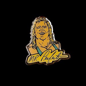 Könnte beinhalten: Anstecknadel mit einer Cartoon-Illustration eines lächelnden Mannes mit langen blonden Haaren und einem türkisfarbenen Oberteil. Der Text "MR. Perfect" ist in gelber Schreibschrift auf der Nadel.