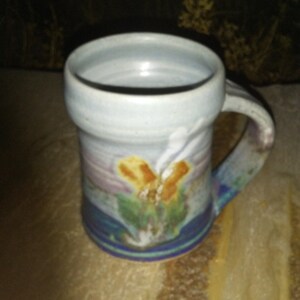 May include: A ceramic mug with a cylindrical body and a thick handle. The exterior features a glaze with blended colors including light blue, purple, green, and a central yellow and white abstract floral-like pattern near the base and middle.