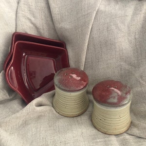 May include: Two small ceramic salt and pepper shakers with a white base and a red top. The shakers are sitting on a beige fabric surface. There are two red ceramic dishes in the background.