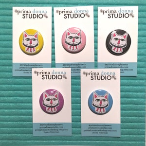 May include: Five 1-inch button pins featuring a cartoon cat wearing a pink collar with a fishbone design. The cat is depicted in five different colors: yellow, pink, black, purple, and blue. Each pin is displayed on a white card with the text "prima donna STUDIO" and social media information.