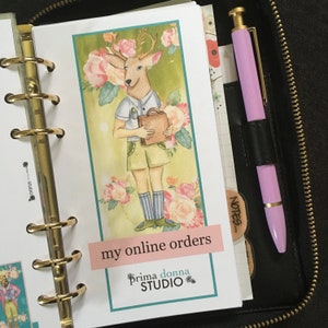 May include: A watercolor illustration of a deer wearing a blue and white shirt and holding a brown briefcase. The deer is standing in a field of pink and white flowers. The image is labeled "my online orders" and "Prima donna STUDIO".