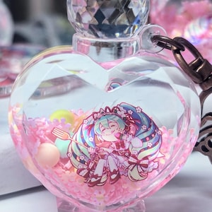 May include: A heart-shaped clear glass bottle charm with a pink glitter and a small anime character inside. The bottle has a crystal-shaped stopper and is attached to a bronze chain.