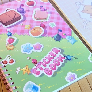 Pu&ograve; includere: Due quaderni a spirale per adesivi. Uno presenta una copertina rosa e verde con adesivi di personaggi dei cartoni animati, cibo e le parole "Sticker Book". L'altro quaderno ha una copertina rosa con un'illustrazione di tre personaggi che dormono in una nuvola.