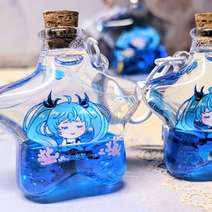 May include: Two clear glass bottles with cork stoppers, each containing blue liquid and a cartoon character illustration. The bottles have a star-shaped design and a metal loop for hanging. The liquid has glitter.