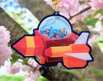 Könnte beinhalten: Ein roter, gelber und weißer Raumschiff-Schlüsselanhänger mit einer durchsichtigen Kuppel, die blaue und orangefarbene Figuren enthält. Der Schlüsselanhänger hängt an einem goldenen Metallclip.