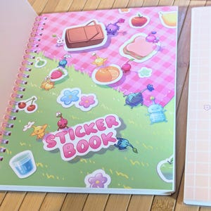 Pu&ograve; includere: Un quaderno di adesivi a spirale con una copertina rosa e verde con illustrazioni di cartoni animati di cibo e creature. Le parole "STICKER BOOK" sono ben visibili. Un secondo quaderno con un motivo a griglia rosa &egrave; visibile.