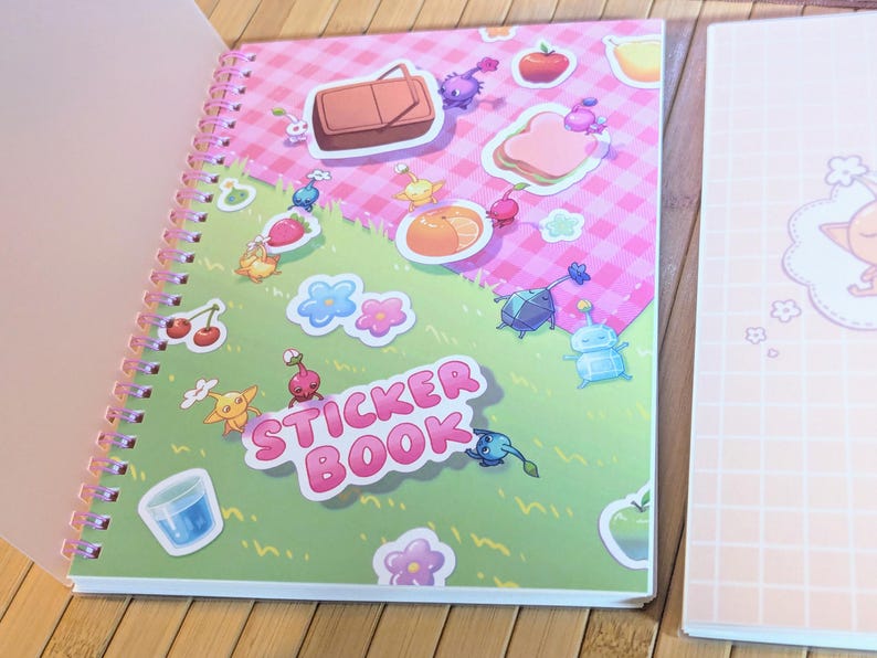 Pu&ograve; includere: Un quaderno di adesivi a spirale con una copertina rosa e verde con illustrazioni di cartoni animati di cibo e creature. Le parole "STICKER BOOK" sono ben visibili. Un secondo quaderno con un motivo a griglia rosa &egrave; visibile.