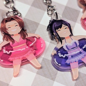 May include: Two acrylic charm keychains featuring anime-style characters in swimsuits and inner tubes. One character wears a pink outfit and the other a purple outfit. Both have smiling faces and are attached to silver keychains.