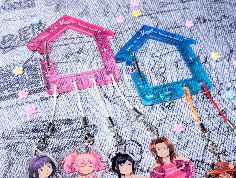 May include: Two clear plastic house-shaped keychains with glitter accents. The keychain on the left is pink and the keychain on the right is blue. Each keychain has a small metal hook and is attached to a white string with a charm at the end. The charms are anime characters.