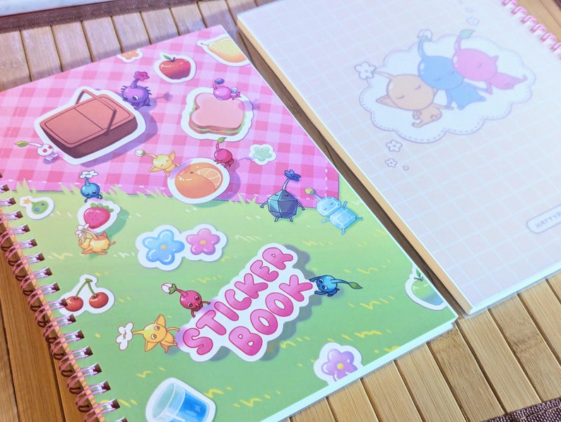 Pu&ograve; includere: Due quaderni a spirale per adesivi. Uno presenta una copertina rosa e verde con adesivi di personaggi dei cartoni animati, cibo e le parole "Sticker Book". L'altro quaderno ha una copertina rosa con un'illustrazione di tre personaggi che dormono in una nuvola.