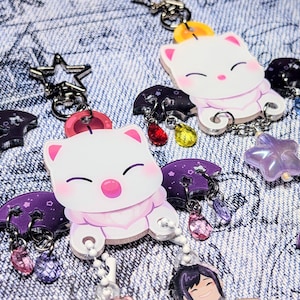 May include: Keychains featuring a white cartoon cat character with pink accents, purple wings, and dangling jewel charms. The keychains have a silver chain and are set against a denim background. Additional charms include stars and other character cutouts.