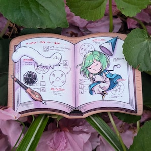 Può includere: Segnalibro in legno con l'illustrazione di un libro aperto con un personaggio dei cartoni animati, una creatura bianca a forma di verme e una penna. Le pagine del libro presentano note e disegni manoscritti. Il segnalibro è circondato da foglie verdi e fiori rosa.