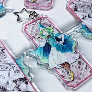 Könnte beinhalten: Ein durchsichtiger Acryl-Schlüsselanhänger mit einer bunten Illustration einer Figur mit grünen Haaren und einem blau-weißen Kleid. Die Figur trägt einen spitzen Hut und hält einen Stab. Der Hintergrund des Schlüsselanhängers ist ein rosafarbenes und weißes Comic-Design.