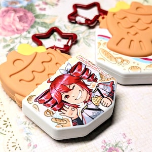 May include: Novelty keyrings with cartoon character and food designs. One has a red star-shaped clasp. The keyrings have a white base with a cartoon illustration and the text "New baguette flavor!!".