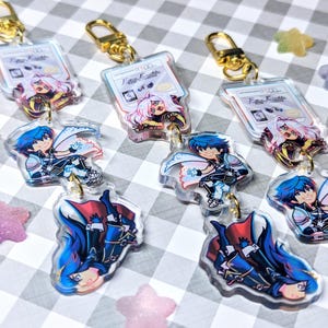 May include: Three clear acrylic keychains featuring a cartoon character with blue hair and a red cape. Each keychain has a smaller charm with a female character with white hair and a blue dress. The charms are attached to a gold chain.