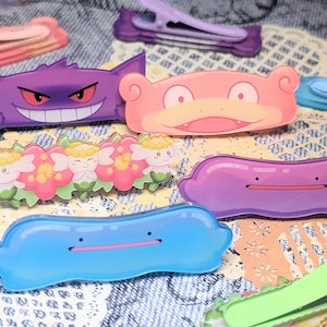 May include: A collection of hair clips featuring various cartoon character designs, including a purple Gengar, a pink Slowpoke, a blue Ditto, a purple Ditto, and a floral pattern with small figures, alongside plain pastel-colored alligator clips.