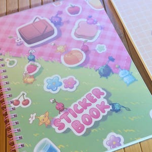 Pu&ograve; includere: Un quaderno a spirale per adesivi con una copertina rosa e verde. La copertina presenta illustrazioni colorate di cibo, fiori e personaggi dei cartoni animati. Le parole "STICKER BOOK" sono stampate in rosa. Un altro quaderno &egrave; visibile sullo sfondo.