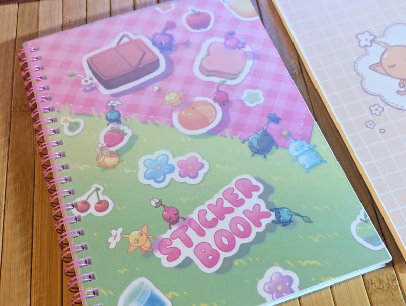 Pu&ograve; includere: Un quaderno a spirale per adesivi con una copertina rosa e verde. La copertina presenta illustrazioni colorate di cibo, fiori e personaggi dei cartoni animati. Le parole "STICKER BOOK" sono stampate in rosa. Un altro quaderno &egrave; visibile sullo sfondo.