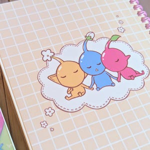 Pu&ograve; includere: Un quaderno a spirale con un motivo a griglia color pesca chiaro. La copertina presenta un'illustrazione di cartoni animati di tre personaggi Pikmin addormentati in arancione, blu e rosa, appoggiati su una nuvola.
