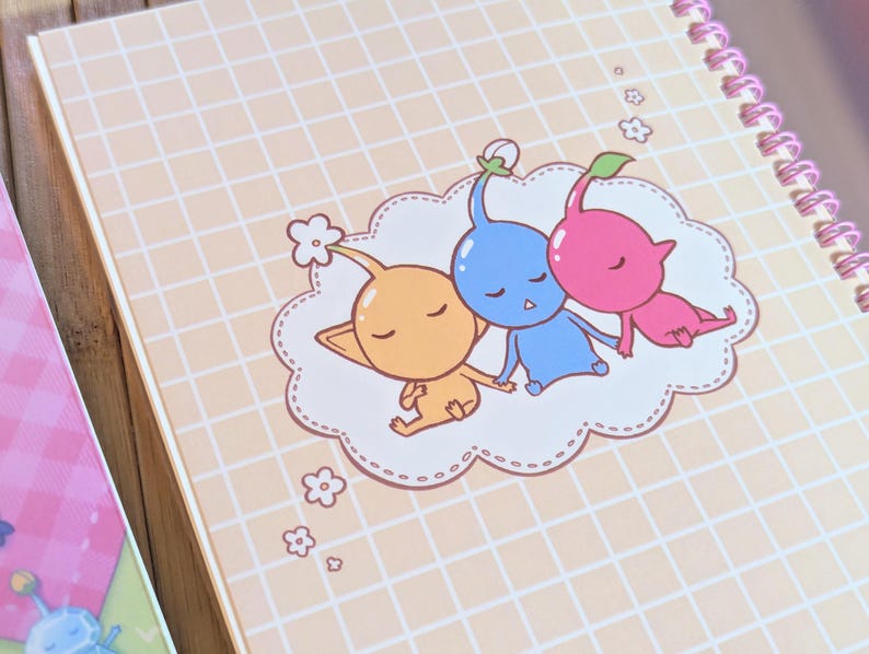 Pu&ograve; includere: Un quaderno a spirale con un motivo a griglia color pesca chiaro. La copertina presenta un'illustrazione di cartoni animati di tre personaggi Pikmin addormentati in arancione, blu e rosa, appoggiati su una nuvola.