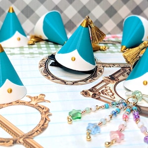 May include: A collection of cone-shaped hair accessories in teal and white, each topped with a gold-colored metal cap and a gold tassel. Some pieces feature a small gold stud, while one includes a dangling charm with multi-colored star beads.