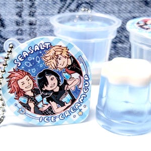 May include: A blue and white Sea Salt Ice Cream Cup keychain with three cartoon characters. The keychain is round with a checkered border. Beside the keychain are several clear plastic cups, one filled with a blue liquid and topped with white foam.