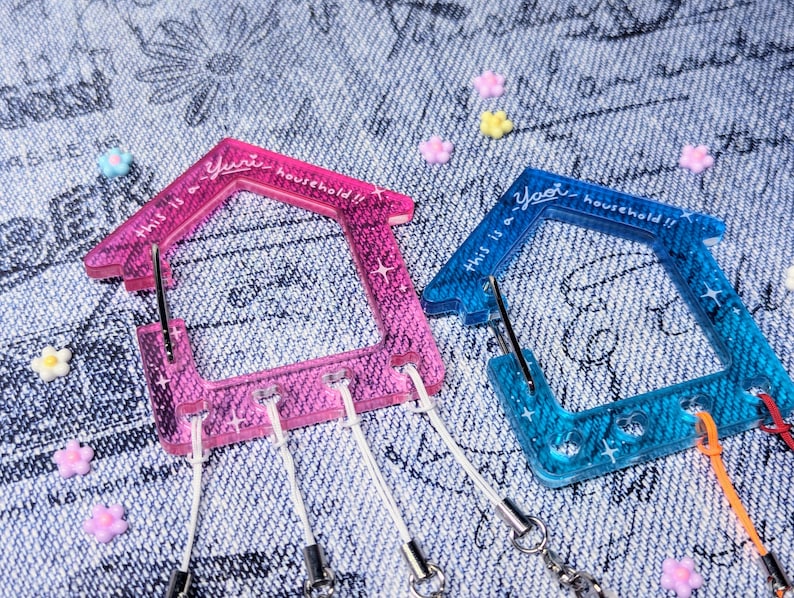May include: Two house-shaped keychains, one pink and one blue, with the text "This is a Yuri household!!" on each. The keychains have sparkly glitter embedded in the plastic.