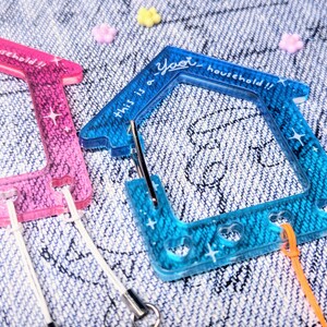 May include: Two colorful plastic keychains shaped like houses with the text "This is a yeet household!!" on the blue keychain. The keychains have glitter embedded in the plastic.