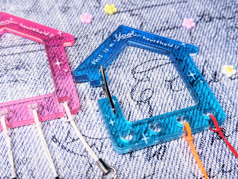 May include: Two colorful plastic keychains shaped like houses with the text "This is a yeet household!!" on the blue keychain. The keychains have glitter embedded in the plastic.