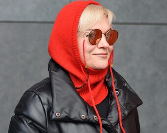 May include: A woman wearing a red knit balaclava with a black leather jacket. The balaclava has a long neck and ties at the back. The woman is wearing sunglasses with a beaded chain.